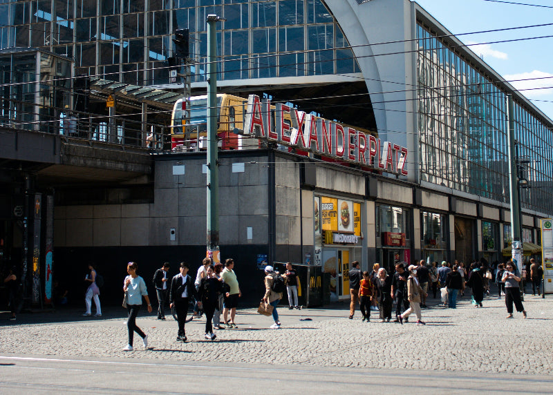 Foto von gut besuchten Alexanderplatz mit der Bahnhofshalle und Berliner S-bahn im Hintergrund. 