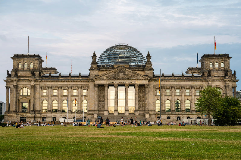 Reichstagsgebäude mit Vorplatz und deutschen Flaggen bei bewölktem Hintergrund. 