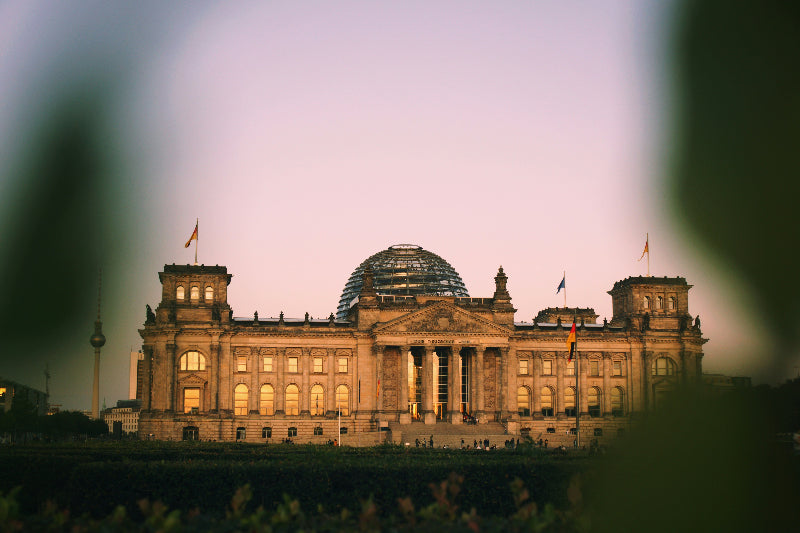 Reichstagsgebäude mit Kuppel und Deutschlandflaggen und Fernsehturm im Hintergrund. 