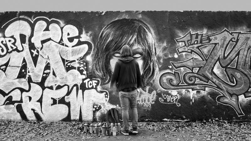 Foto von einem Künstler vor einer Graffiti wand. Auf dem Boden stehen Sprühdosen. 